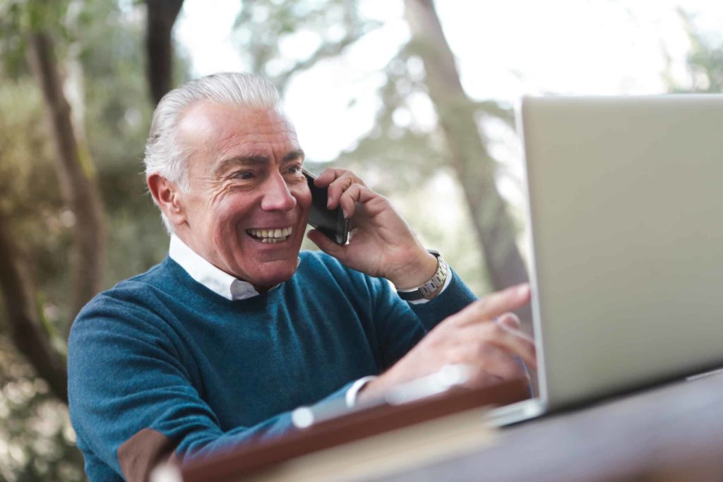 Canva Selective Focus Photo of Smiling Elderly Man in Blue Sweater Sitting by the Table Talking on the Phone While Using a Laptop scaled 1 for