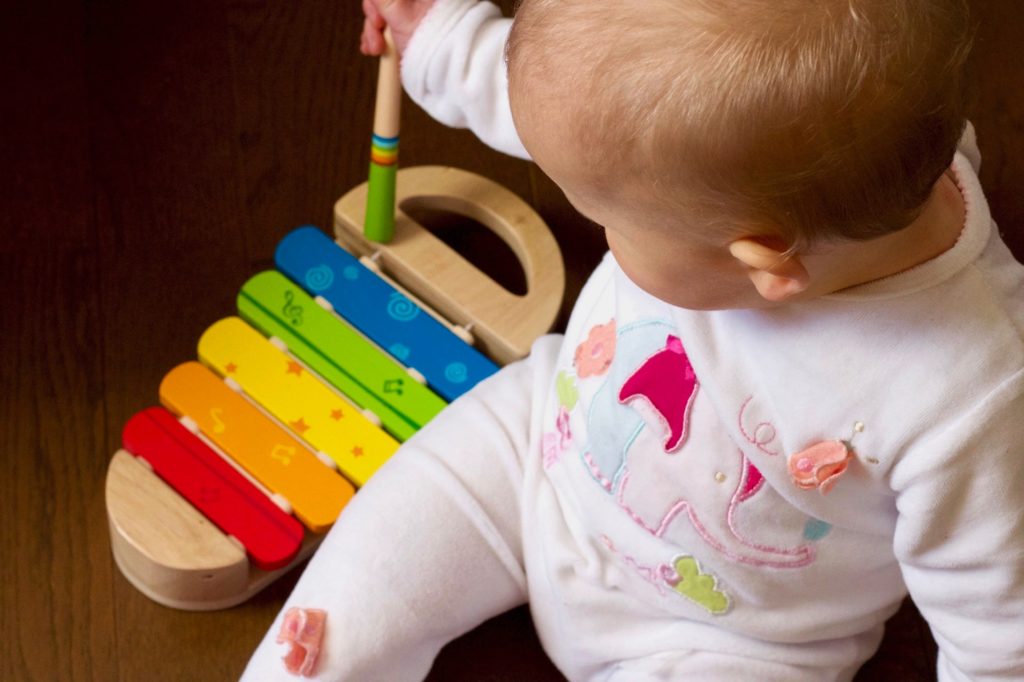 Baby of about eight months wearing a onesie with an elephant on the front and playing with a xylophone.