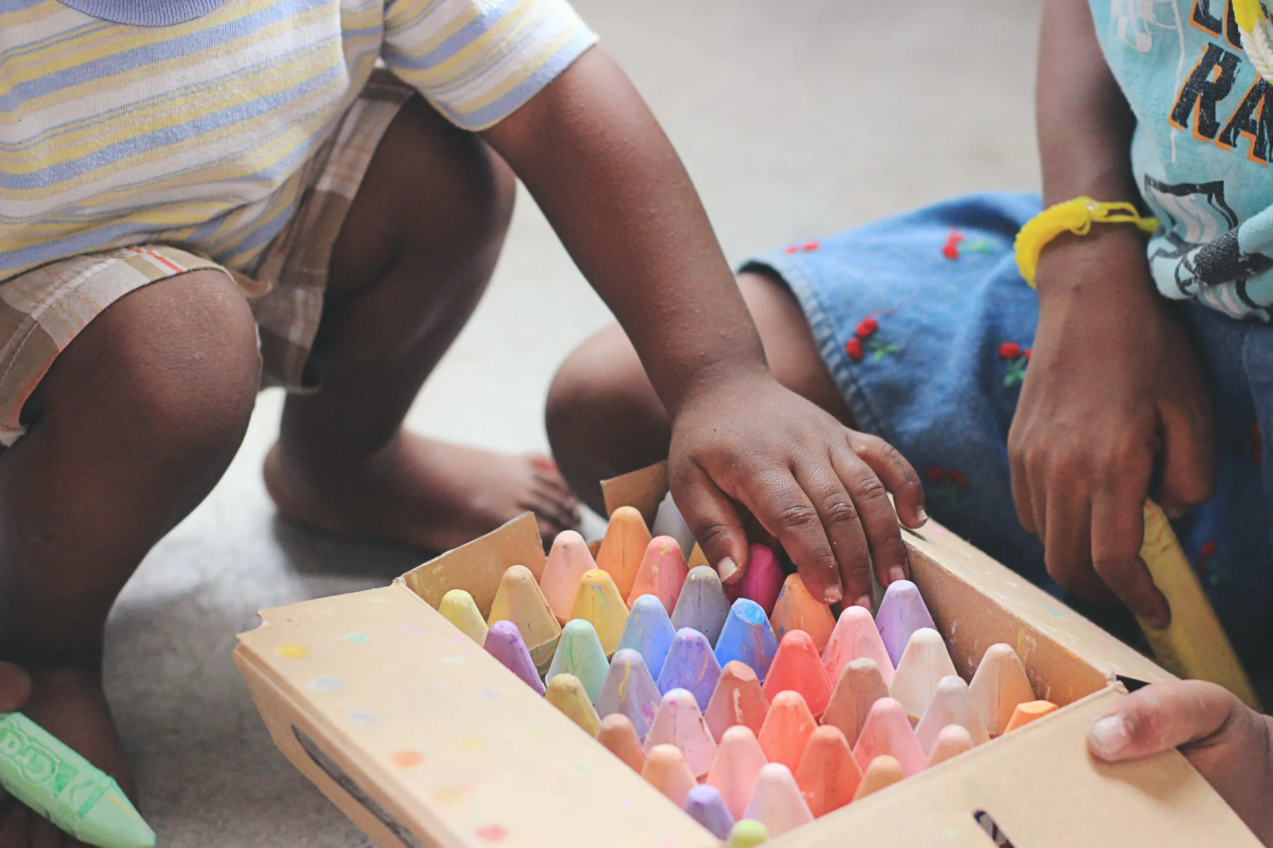 children at a head start program. Only their hands and lower half of their bodies are visible as they pick out pieces of multicolored chalk from a box on the ground. 