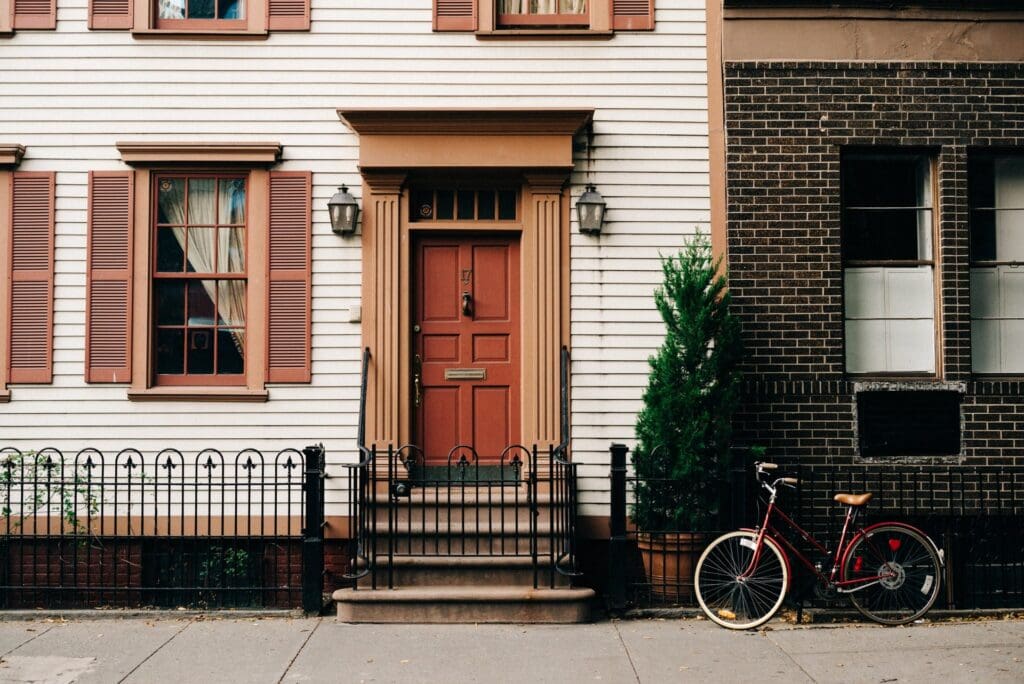 Door of a white house with brown trim and shutters. A bicycle leans against the railing outside. Cover image in article on how to get free housing.