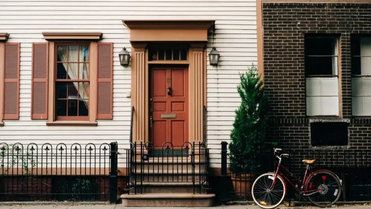 Door of a white house with brown trim and shutters. A bicycle leans against the railing outside. Cover image in article on how to get free housing.