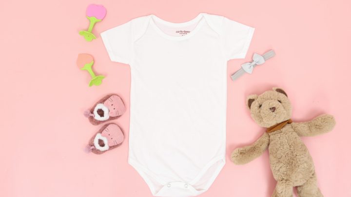 White baby onesie on pink background surrounded by teddy bear, baby shoes, and hair bows in article about free stuff for single moms