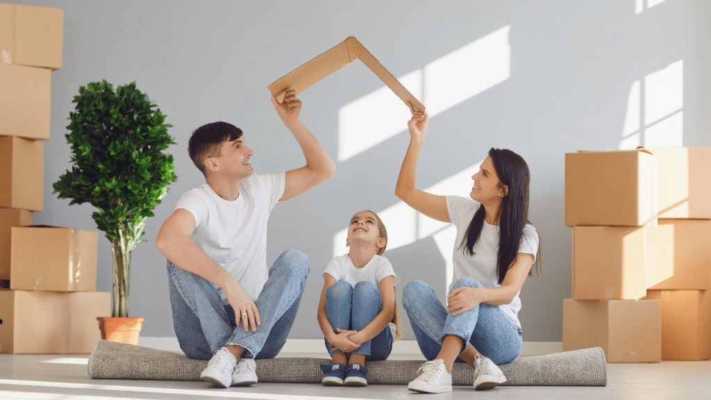 couple holds cardboard house cutout over their children while moving in, surrounded by moving boxes, after finding low income housing with no waiting list