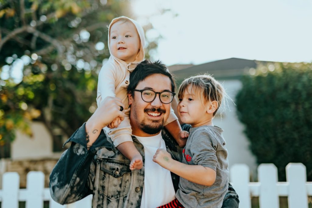 By Unsplash user Nathan Dumlao. A  smiling man stands in front of a house with two children - one on his shoulders, the other cradled in his arm. They have their home through alternative options and didn't have to worry about finding  low income housing with no waiting list.