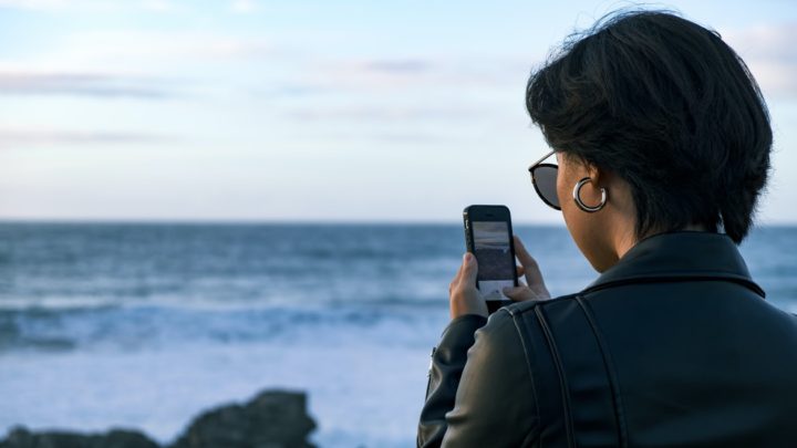 By Unsplash user Portuguese Gravity. A short haired woman wearing sunglasses faces the ocean while on her phone. Maybe she is using the Be My Eyes app to describe the color of the waves.