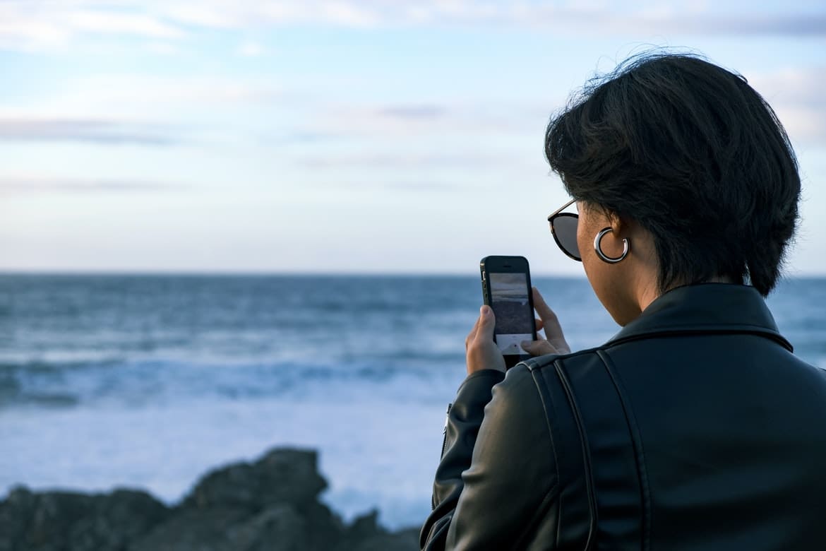 By Unsplash user Portuguese Gravity. A short haired woman wearing sunglasses faces the ocean while on her phone. Maybe she is using the Be My Eyes app to describe the color of the waves.