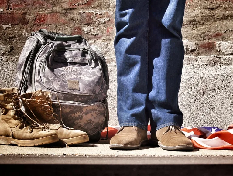 By Unsplash user Benjamin Faust. A veteran, presumably male due to dress, stands by an American flag, combat boots, and a combat backpack. Now that he is home, he should start looking for Florida veterans benefits.