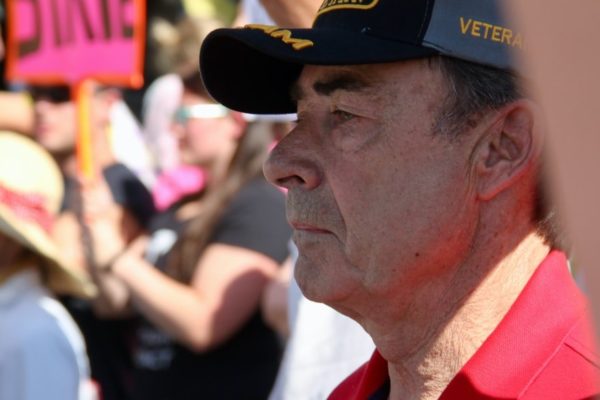 By Unsplash user Manny Becerra. A male Vietnam veteran solemnly stares off while in a crowd of people holding signs.