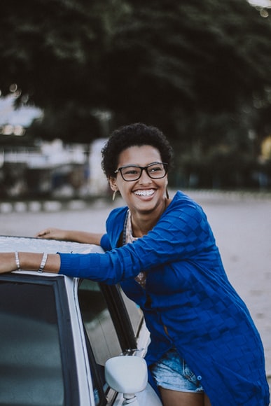 By Unsplash user Caique Silva. A woman in a blue shirt with black glasses smiles as she leans on her white vehicle. She's successfully received assistance through the Arizona Voluntary Vehicle Repair Program.