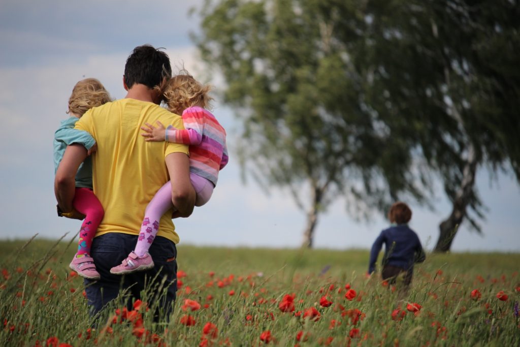 father carrying twins and child running ahead of them in a field in article on Colorado Works