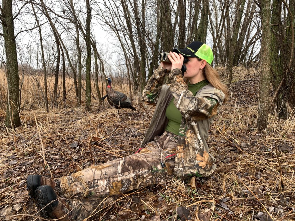 woman sitting in the woods looking through binoculars on a turkey hunt in article on idaho veterans discounts