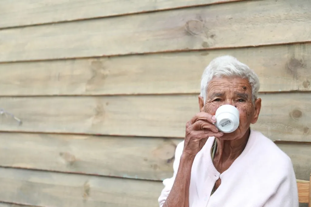 elderly man in a white shirt with white hair sipping coffee against a wood paneled wall in article on adult public assistance