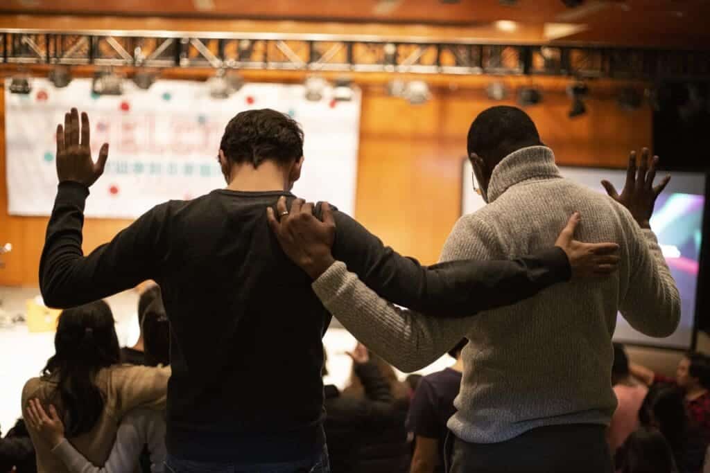 A group prays together at Love INCBy Unsplash user Sam Balye. Two men have their arms around each other as the other hands are lifted in prayer.