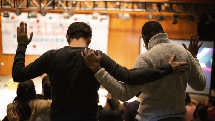 By Unsplash user Sam Balye. Two men have their arms around each other as the other hands are lifted in prayer.