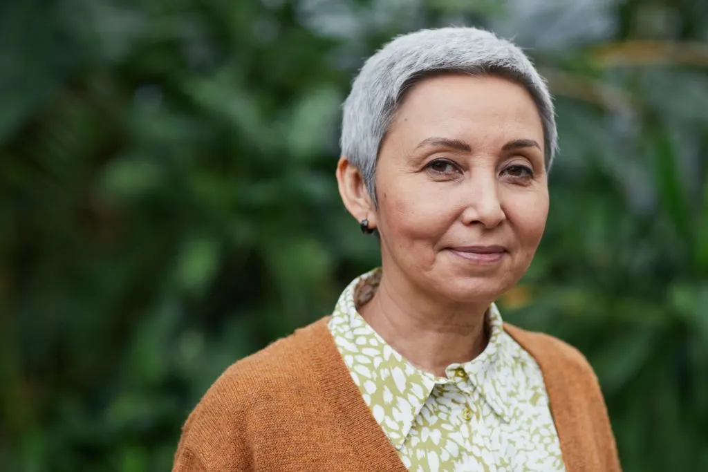 A senior woman with short cropped gray hair wearing an orange sweater and looking into the camera in article on senior benefits in Alaska