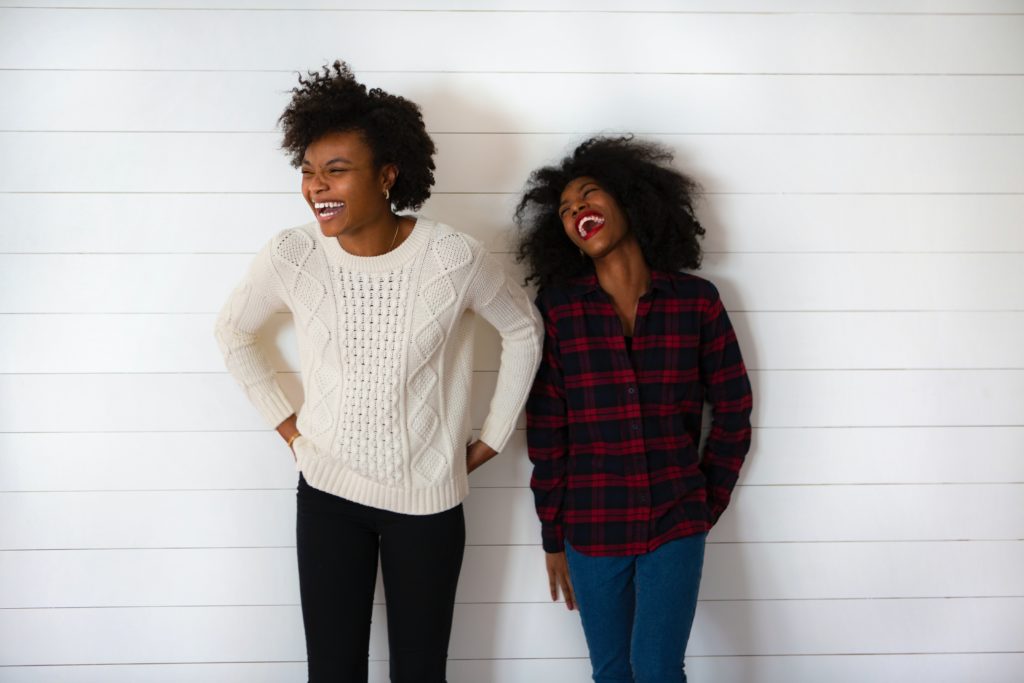 two women laughing together against a white brick wall in article on what is buy nothing