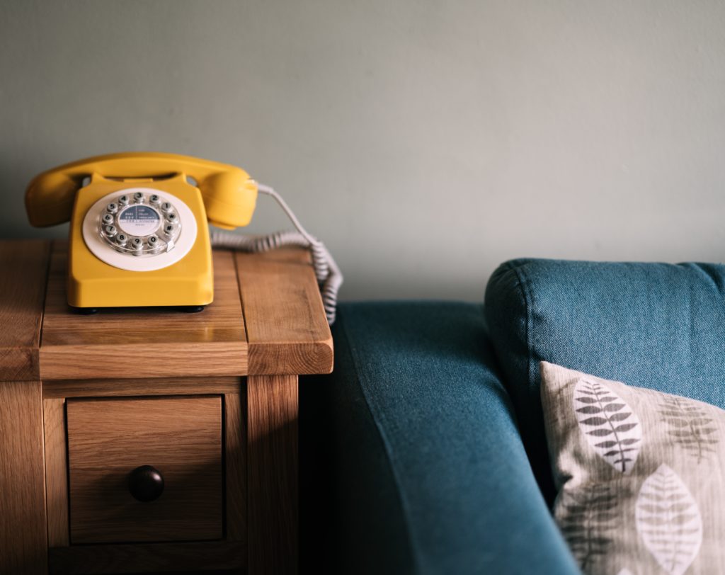 yellow rotary phone on end table next to blue couch in article on cheap landline phone service for seniors