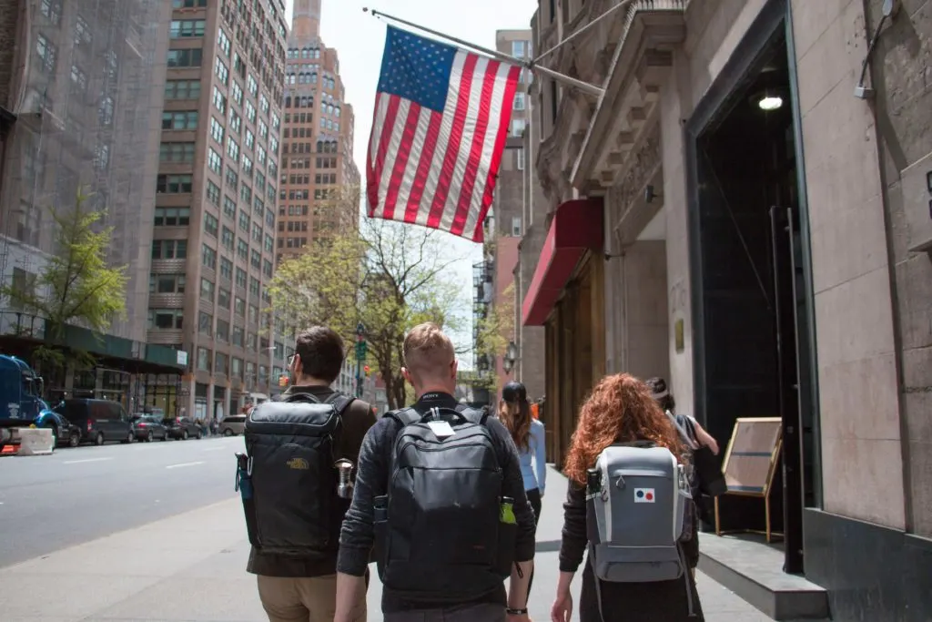college students with backpacks walking down new york city street toward american flag in article on how do immigrants become citizens