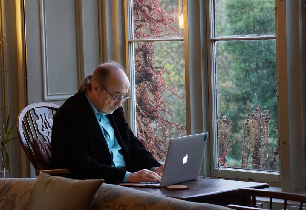 elderly man sitting in front of laptop computer next to a window in article on how to find a social worker for elderly