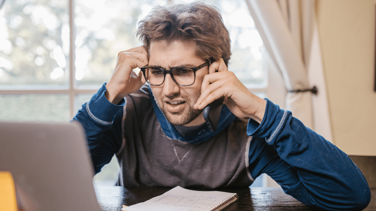 man wearing glasses looks at his laptop confused while holding his phone to his ear. He is trying to ask someone if EBTEdge is legit