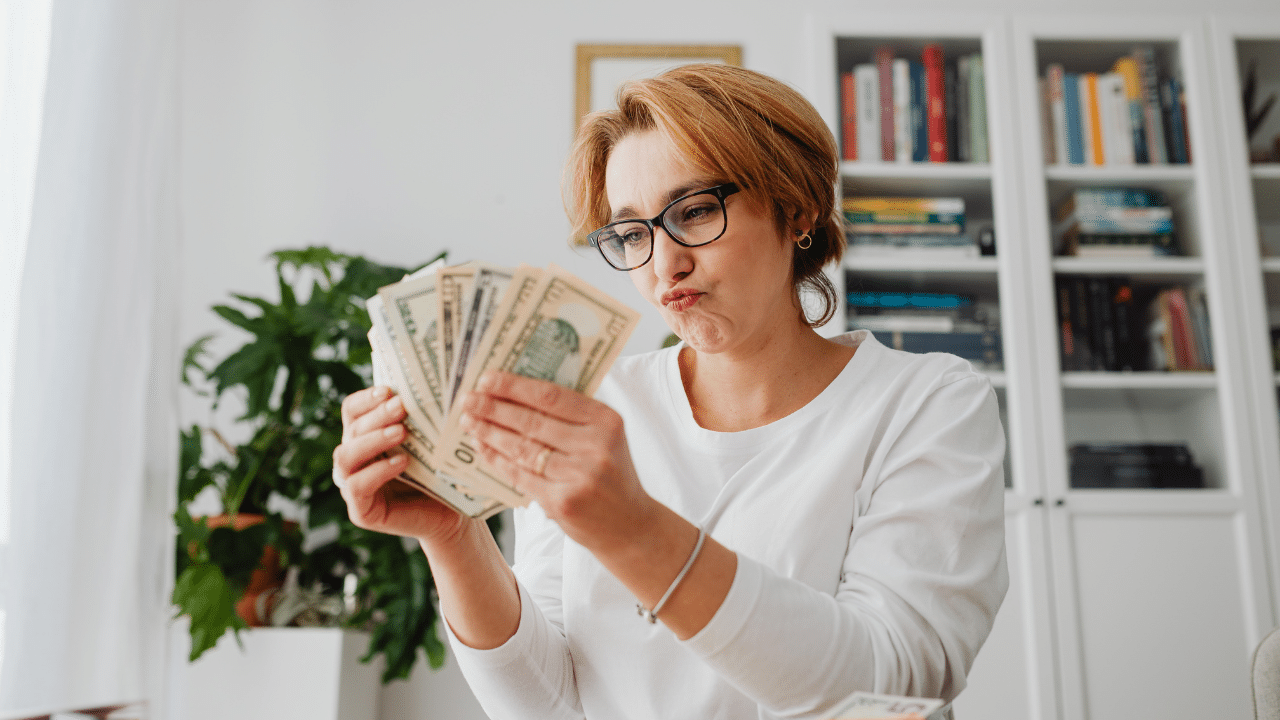 A woman with glasses counts a stack of U.S. dollar bills at home, looking thoughtful. She is seated in front of a bookshelf and a houseplant, wearing a white shirt, after reading about how to get free money in April 2025.