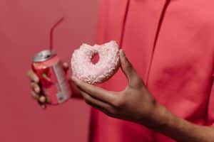 Close-up of a hand holding a pink doughnut and a soda can against a pink backdrop.