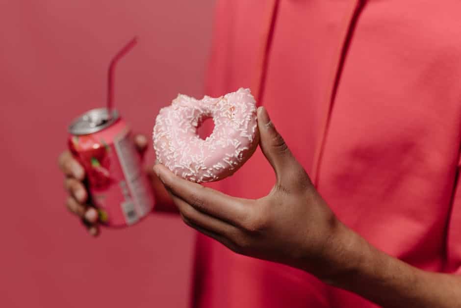 Close-up of a hand holding a pink doughnut and a soda can against a pink backdrop.